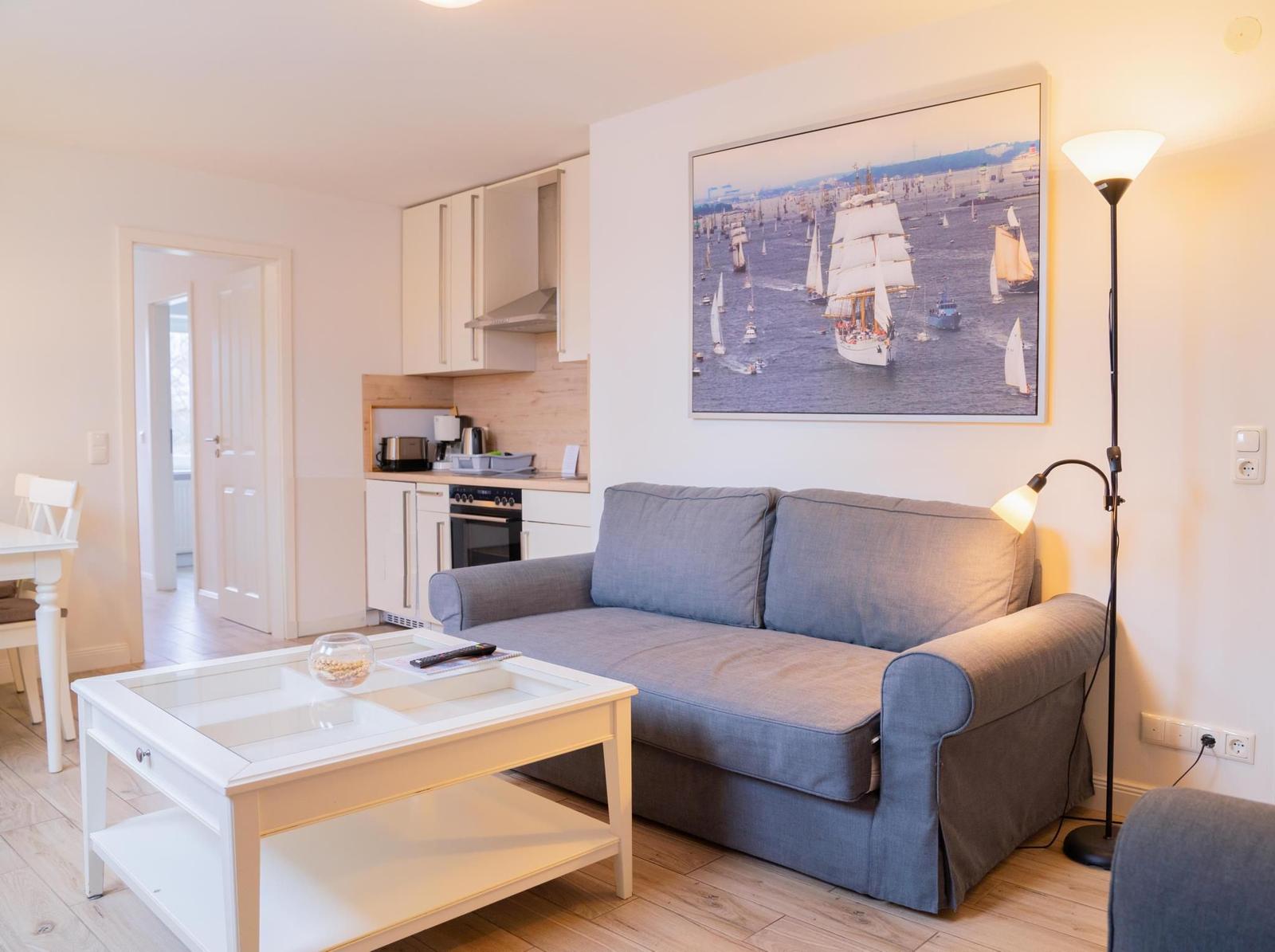 Wohnzimmer mit Sofa, Tisch und Küchenbereich. Wandbild mit Segelbooten.
