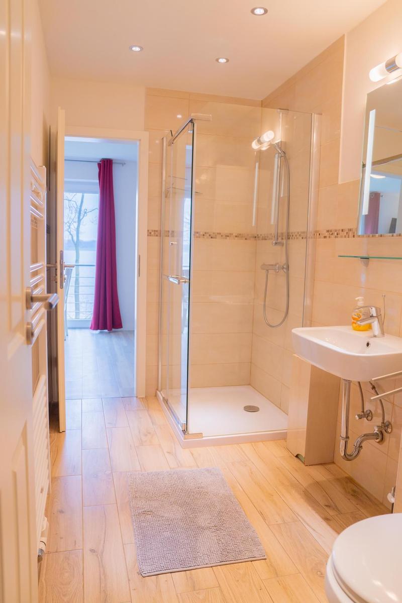 Badezimmer mit Dusche, Waschbecken und Toilette. Holzfußboden und Tür zum Balkon.