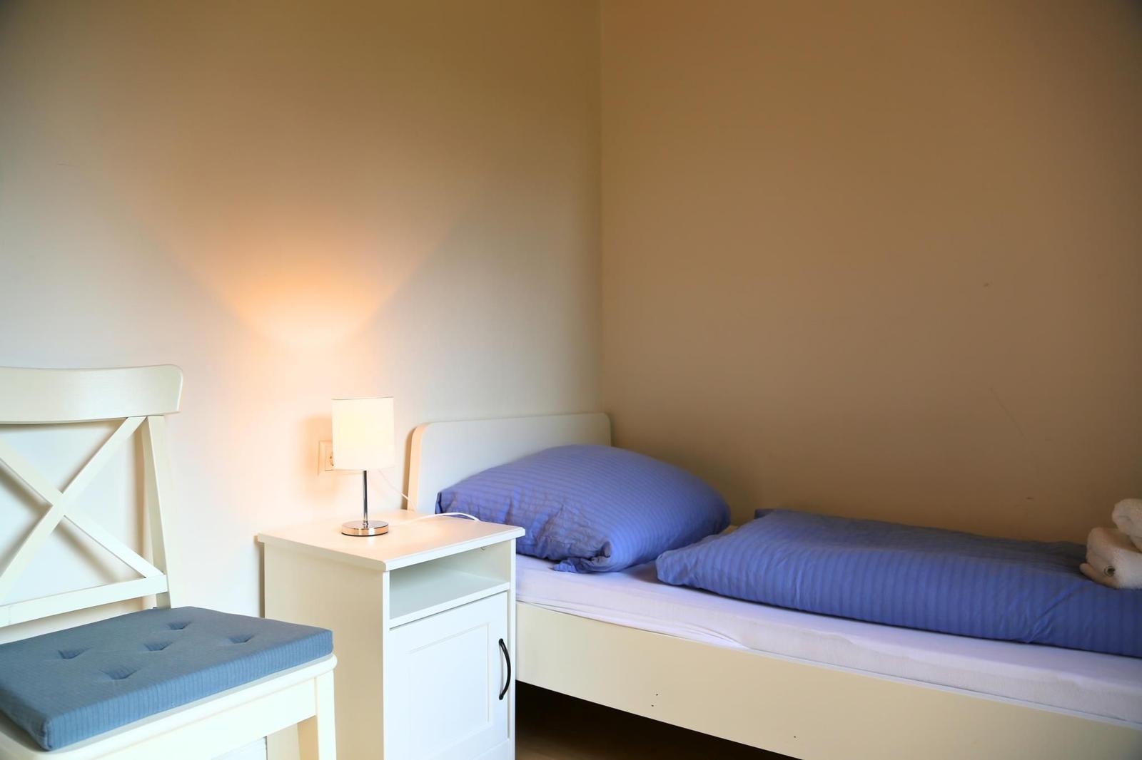 Ein Bett mit blauer Bettwäsche, Nachttisch und Stuhl in einem Raum mit hellen Wänden.