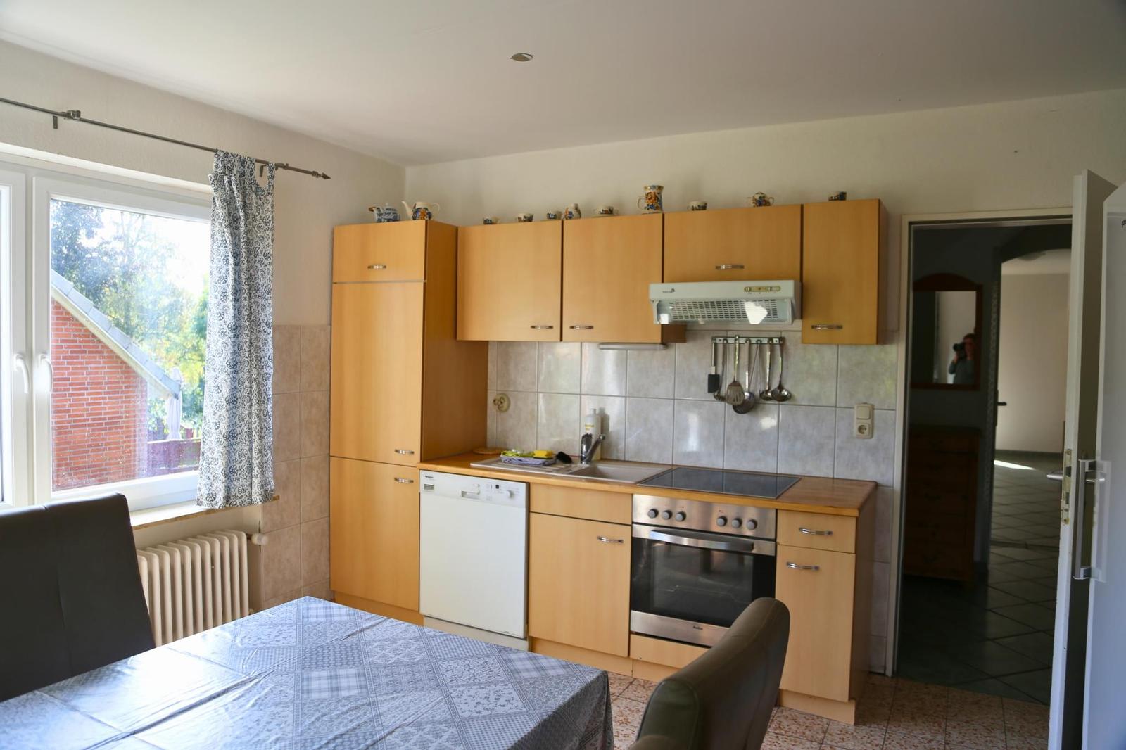 Küche mit Holzschränken, Spüle, Herd und Dunstabzug. Tisch mit Tischdecke und Fenster mit Vorhang.