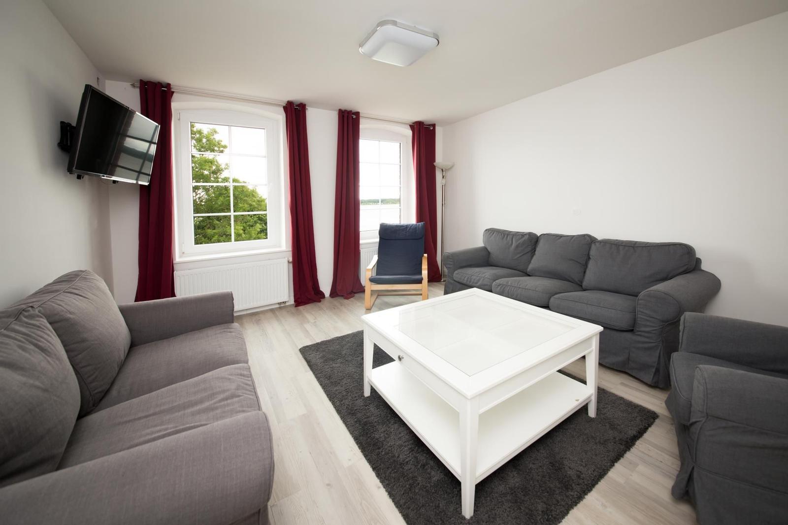 Wohnzimmer mit grauen Sofas, weißem Tisch und roten Vorhängen an den Fenstern.