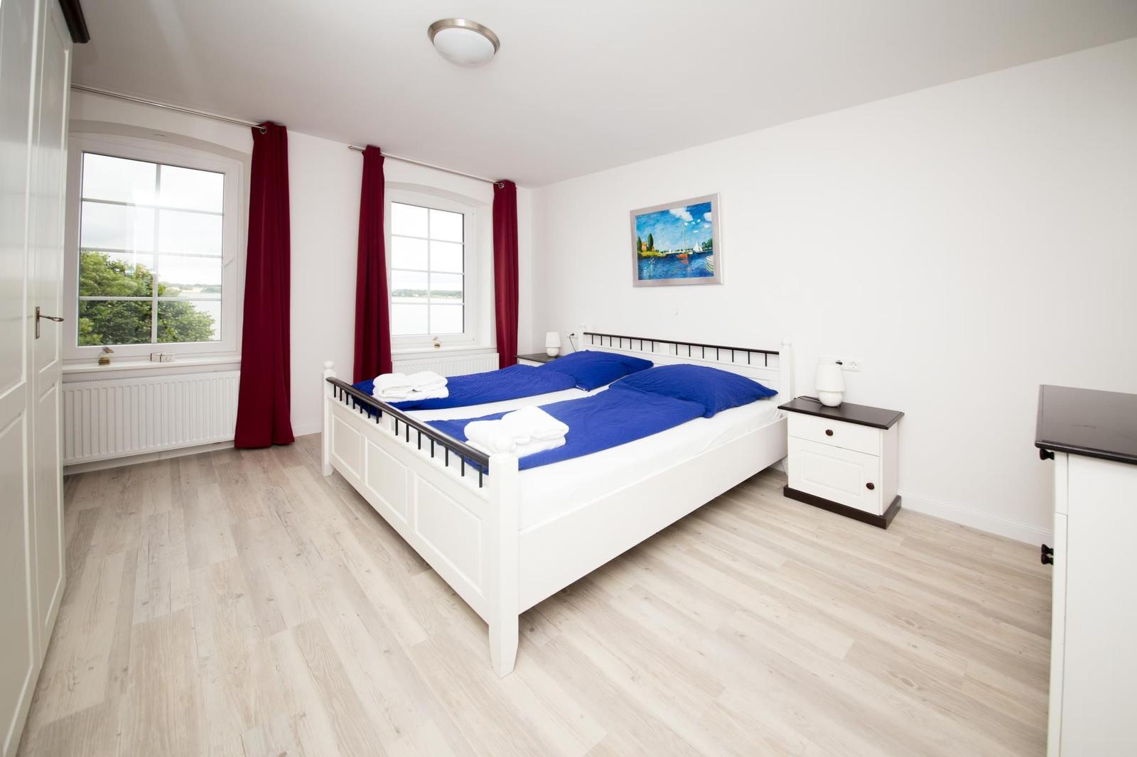 Schlafzimmer mit Doppelbett, zwei Fenstern und roten Vorhängen.