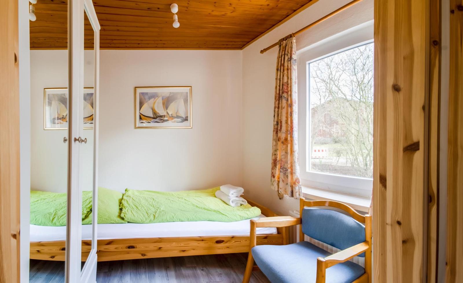 Ein Schlafzimmer mit Bett, Stuhl und Fenster. Holzdecke und Kleidung sind sichtbar.