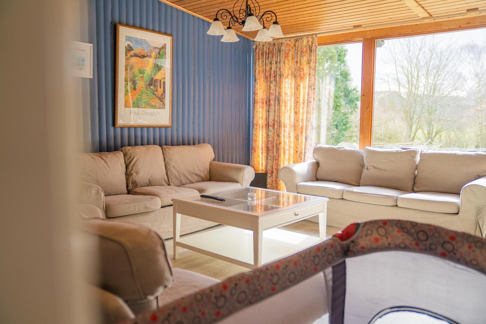Wohnzimmer mit zwei Sofas, Glas-Tisch und blauer Wand. Große Fenster mit Blick auf Außenbereich.