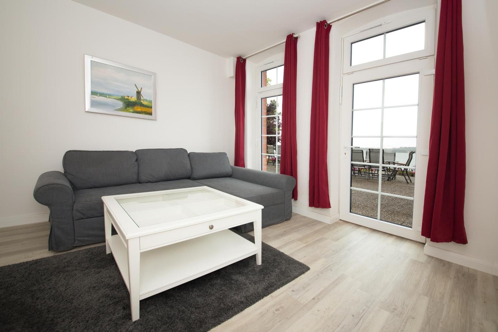 Wohnzimmer mit grauem Sofa, weißem Couchtisch und roten Vorhängen an der Glastür.