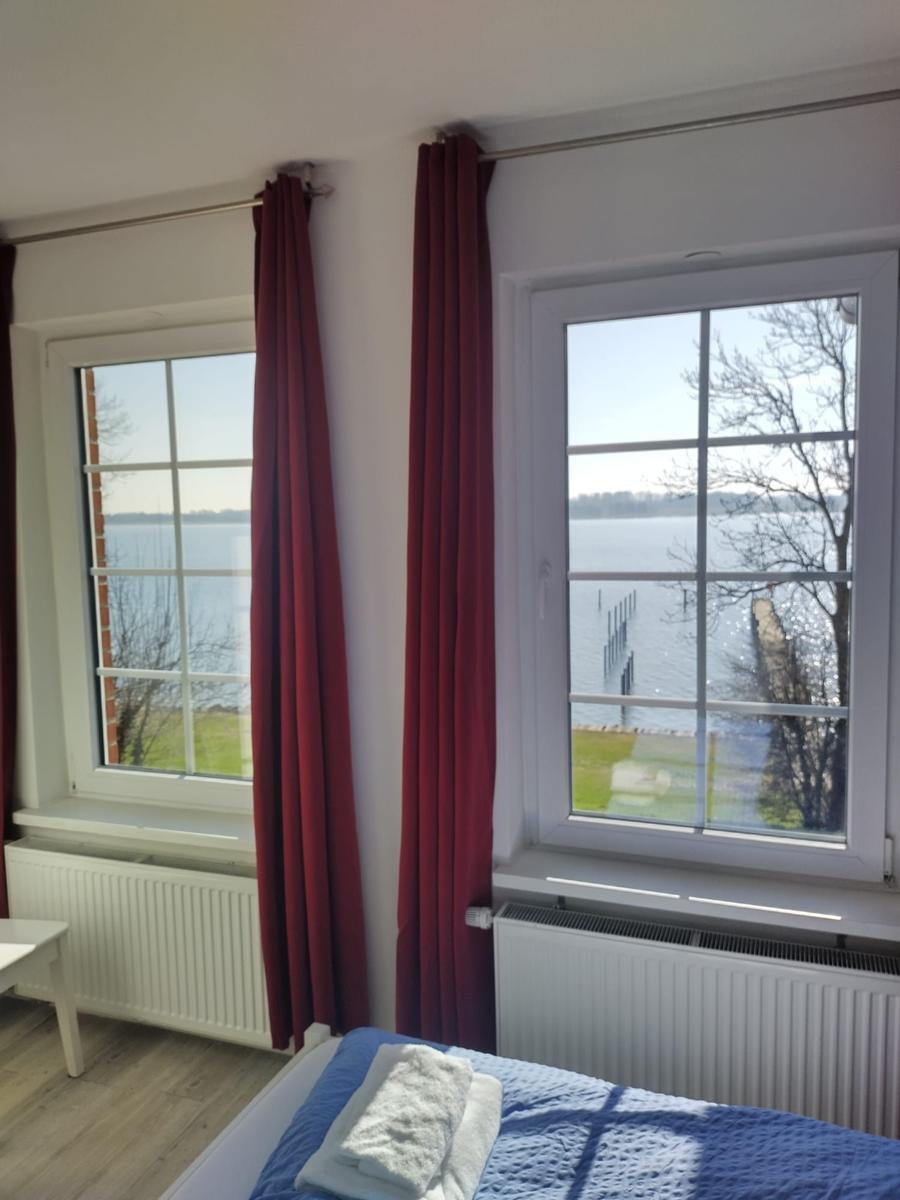 Zimmer mit Blick auf Wasser, zwei Fenster, rote Vorhänge, Bett mit blauer Bettwäsche.