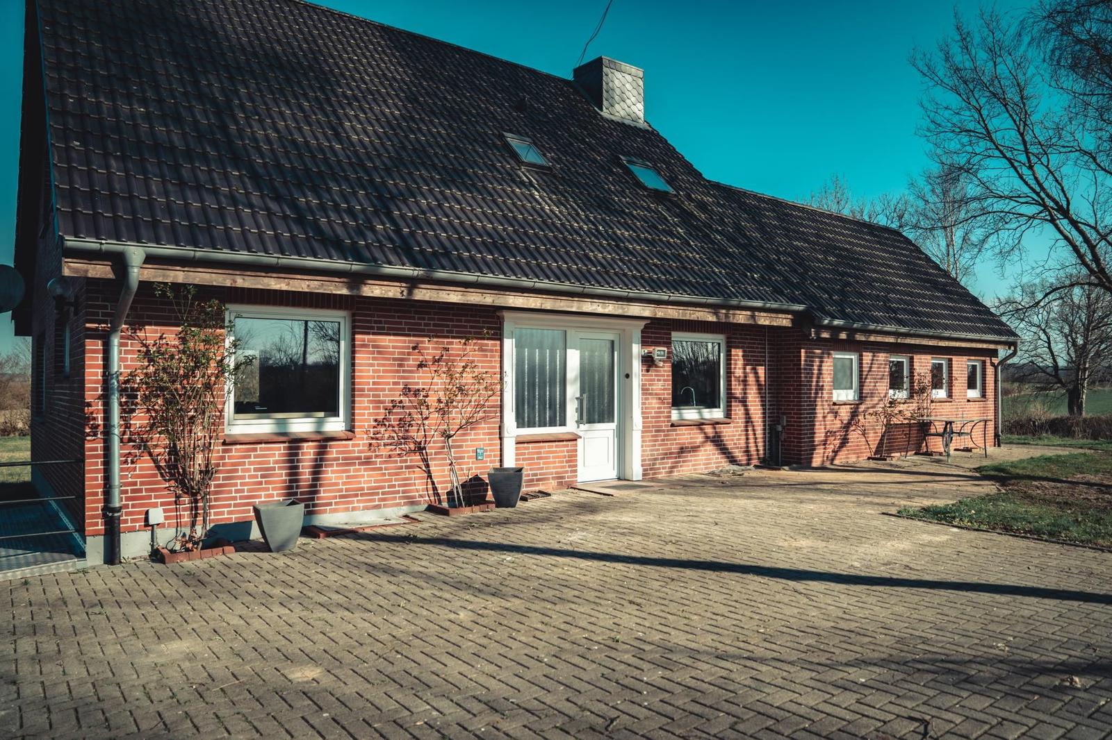 Rotes Backsteingebäude mit dunklem Dach und weißen Fensterrahmen. Vor dem Haus befindet sich ein gepflasterter Hof.