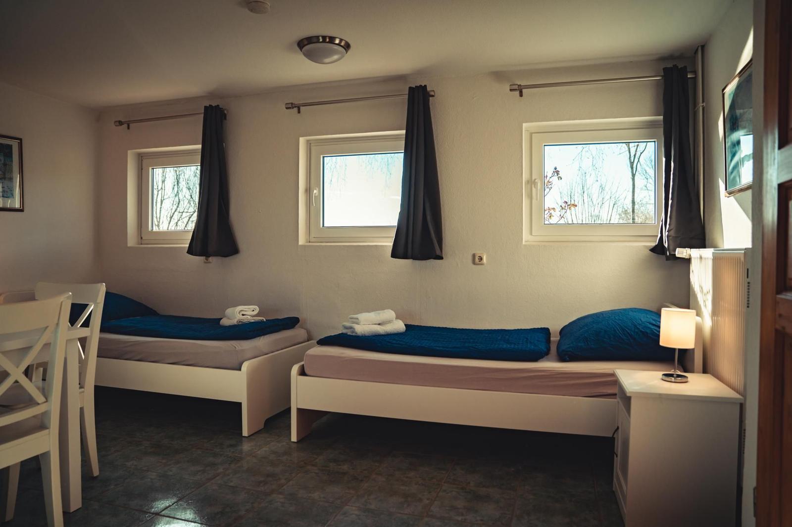 Zwei Betten mit blauen Decken, drei Fenster mit dunklen Vorhängen und ein Nachttisch mit Lampe.