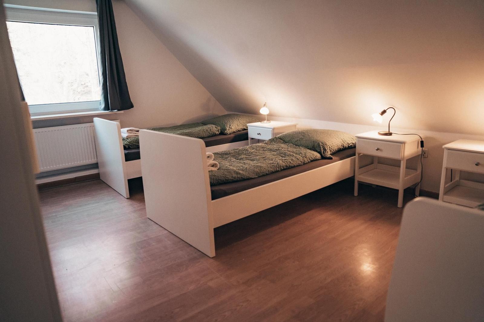 Zwei weiße Betten mit grünen Decken stehen unter einem Dachschrägen. Drei Nachttische mit Lampen.