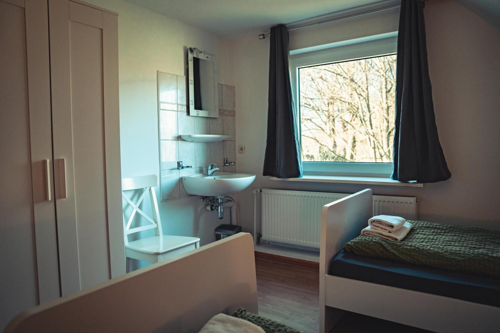 Zimmer mit Bett, Waschbecken und Fenster mit Blick auf Bäume.