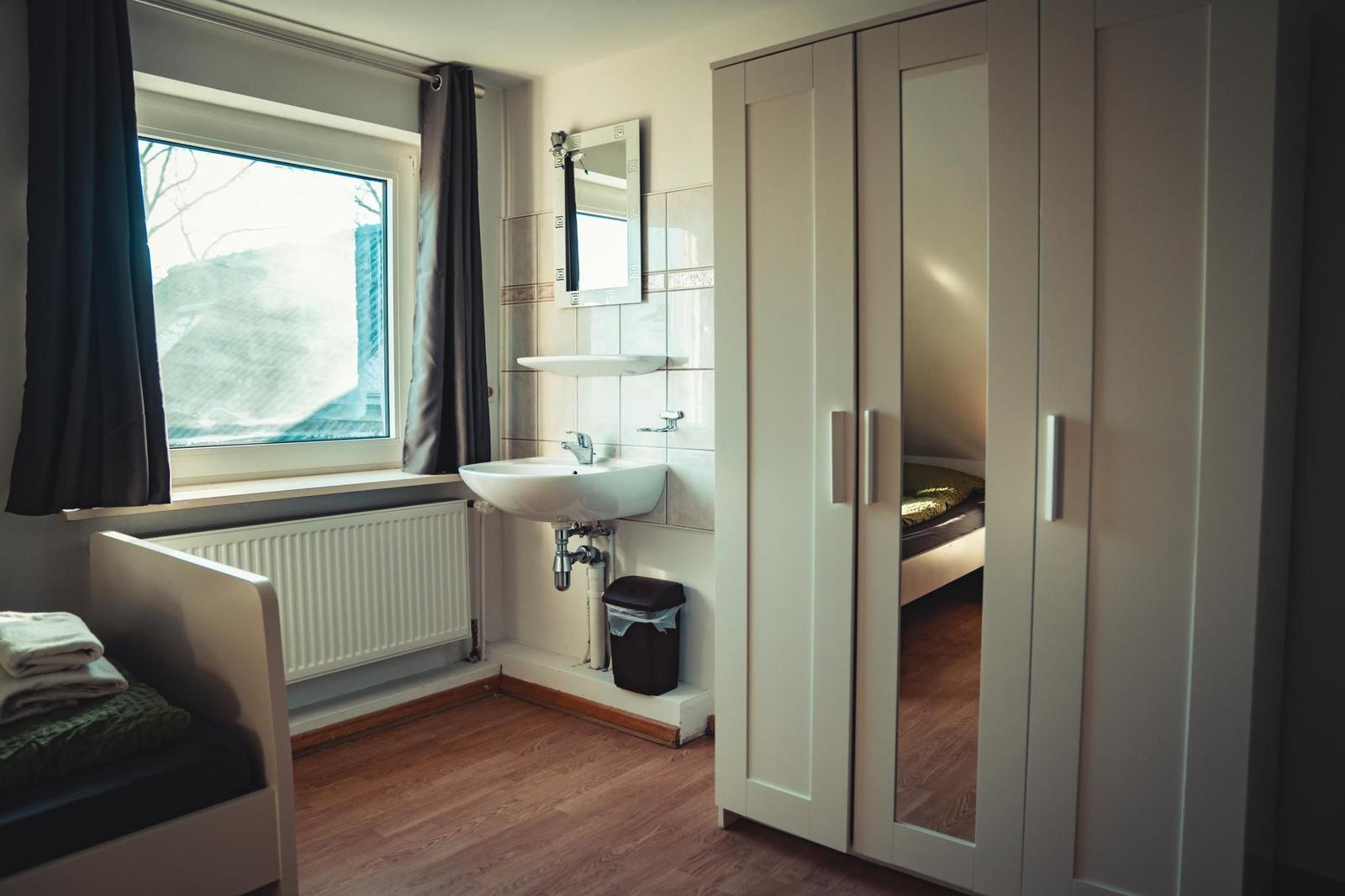 Schlafzimmer mit Fenster, Waschbecken und Kleiderschrank. Spiegel und Heizkörper sind sichtbar.