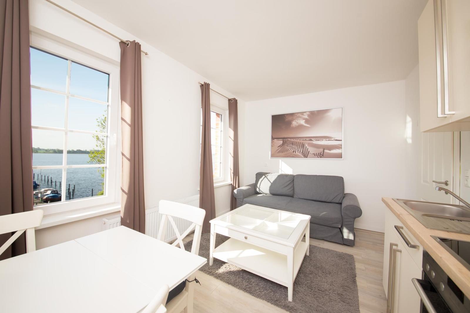 Wohnung mit Küchenzeile, Sofa und Tisch. Blick auf Wasser durch Fenster.
