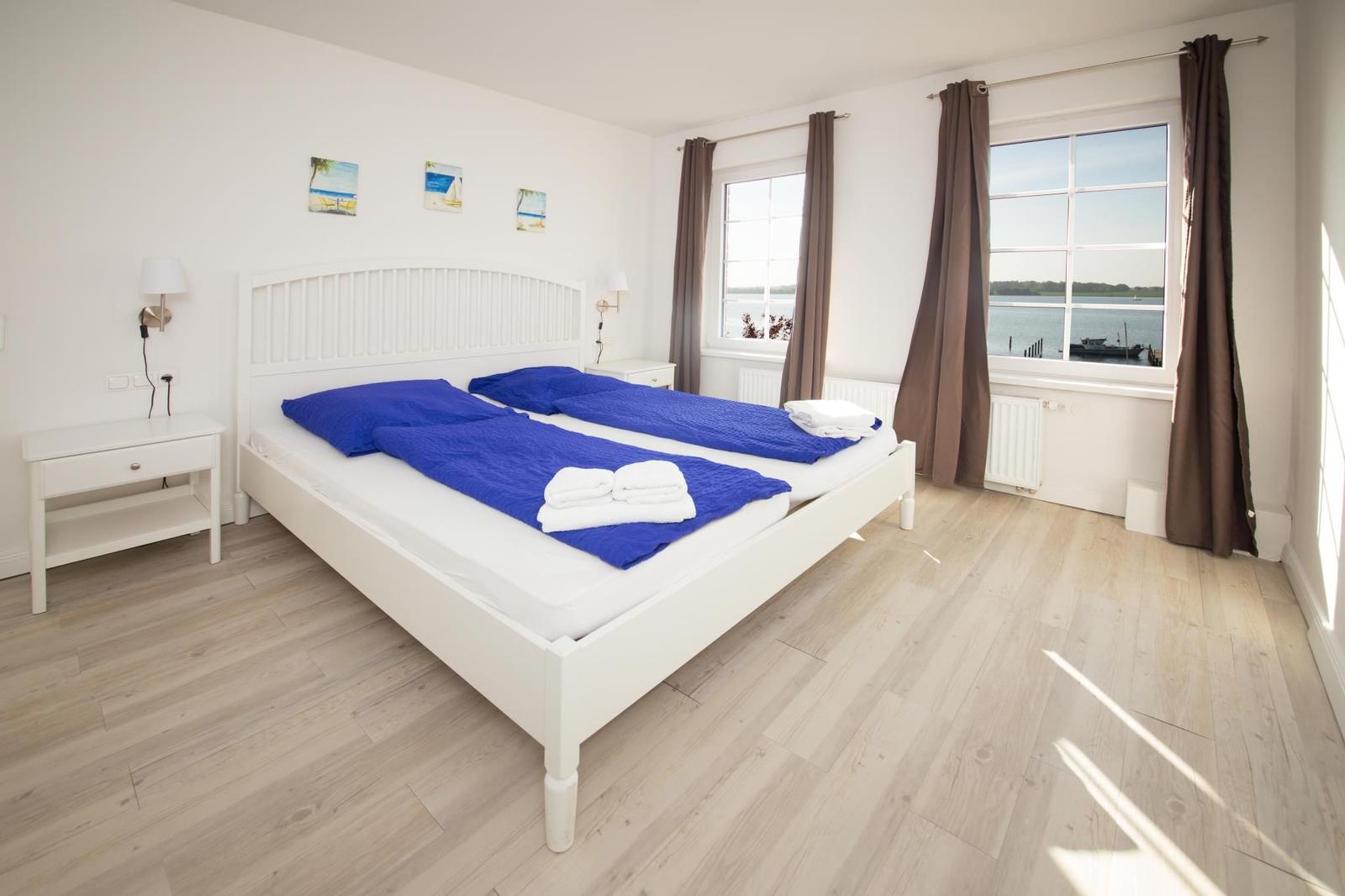 Schlafzimmer mit weißem Bett, blauen Bettwäsche und Blick auf Wasser.