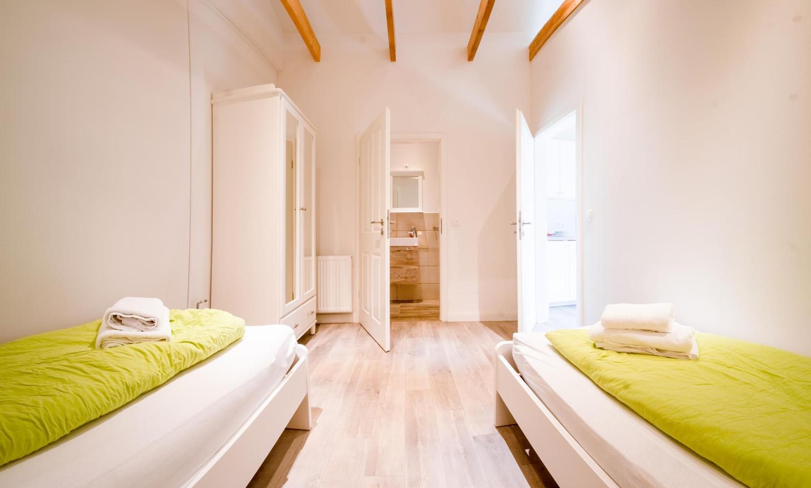 Zwei Betten mit grünen Decken, Schrank und Blick in Bad und Küche.