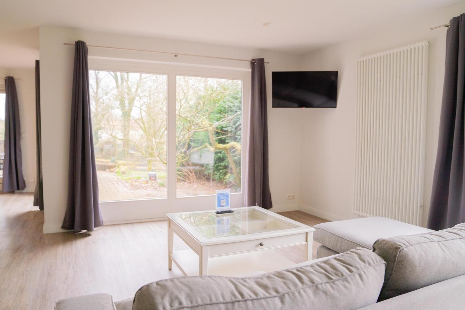Wohnzimmer mit Sofa, Glas-Tisch und Fernseher an der Wand. Große Fenster mit Blick auf Garten.