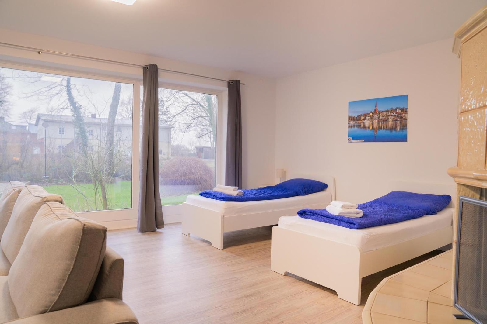 Zwei Betten mit blauen Decken, Sofa und großes Fenster mit Blick auf Garten.
