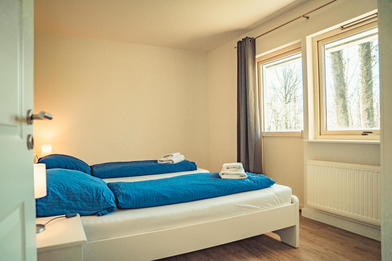 Schlafzimmer mit Doppelbett, blauen Bettwäsche und Fenster mit Blick auf Bäume.