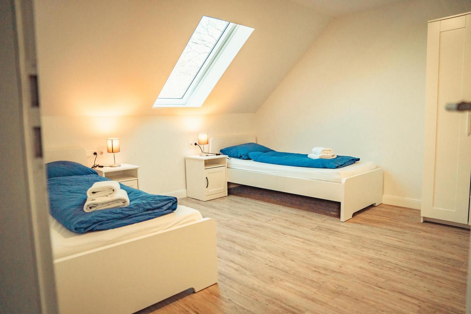 Zwei Betten mit blauen Bettwäschen und Nachttischen unter einem Dachfenster.