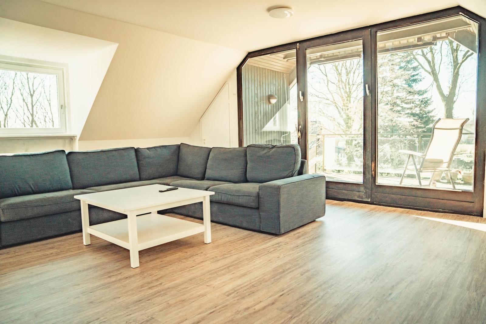 Wohnzimmer mit grauem Sofa, weißem Tisch und Holzboden. Große Glasfront mit Blick auf Balkon.