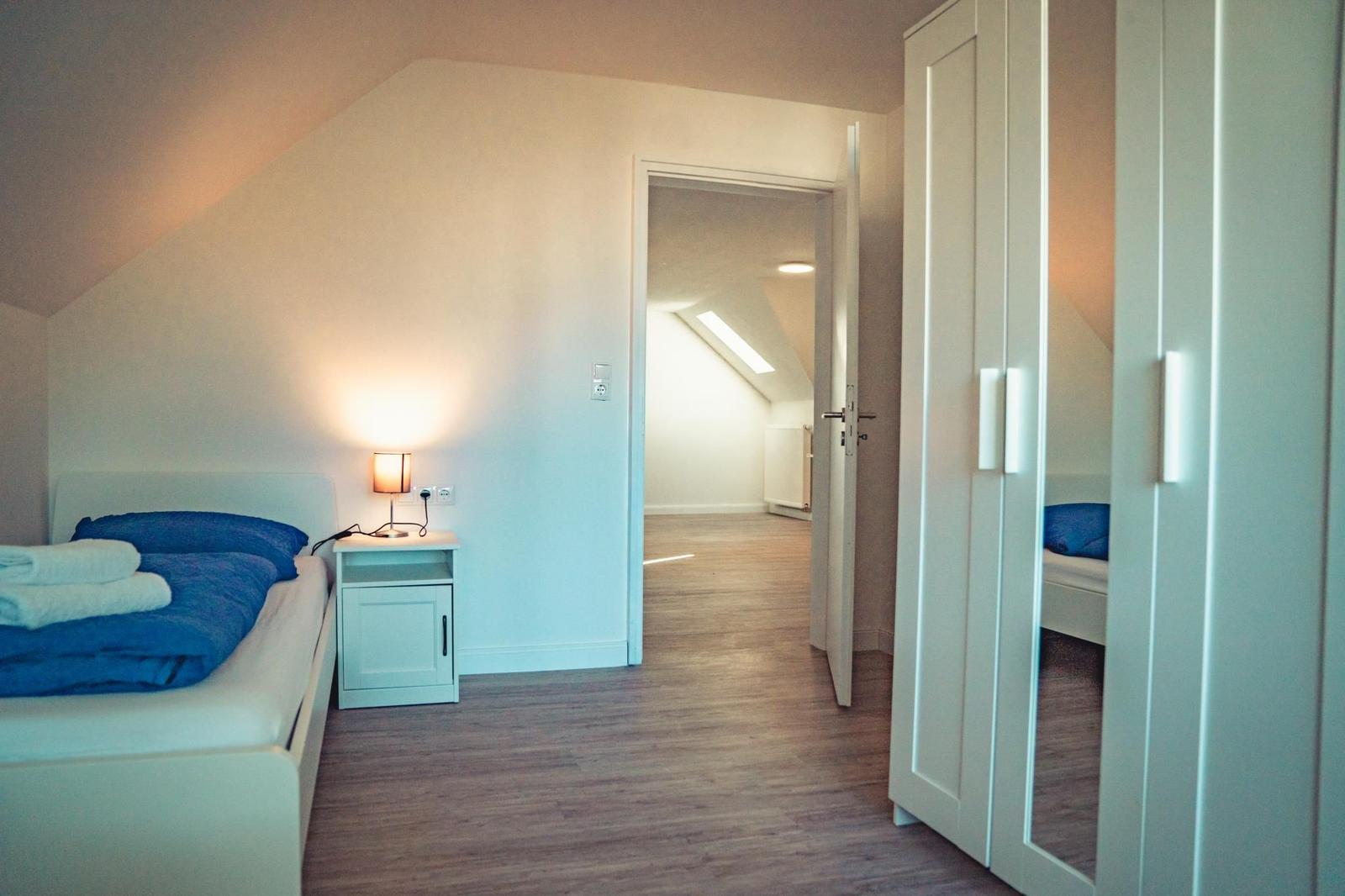 Schlafzimmer mit Bett, Nachttisch und Kleiderschrank. Tür führt in einen Flur mit Dachschrägen.