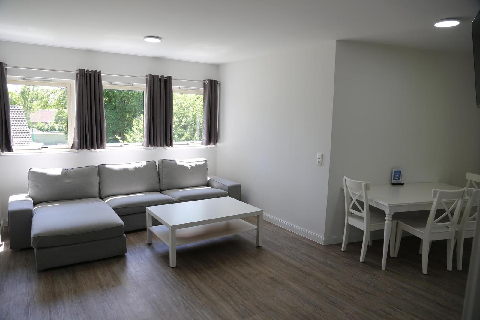Wohn- und Essbereich mit Sofa, Tisch und Stühlen. Fenster mit Vorhängen und Holzboden.