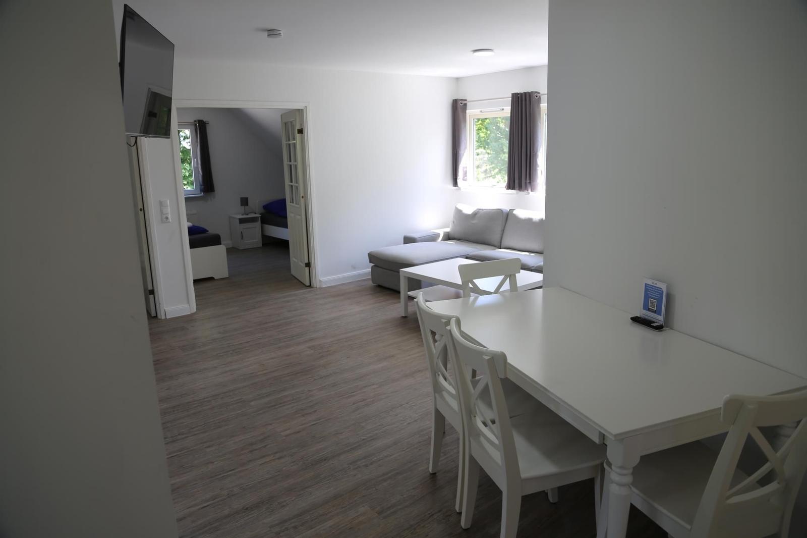 Wohn- und Essbereich mit Sofa, Tisch und Stühlen. Im Hintergrund ist ein Schlafzimmer sichtbar.