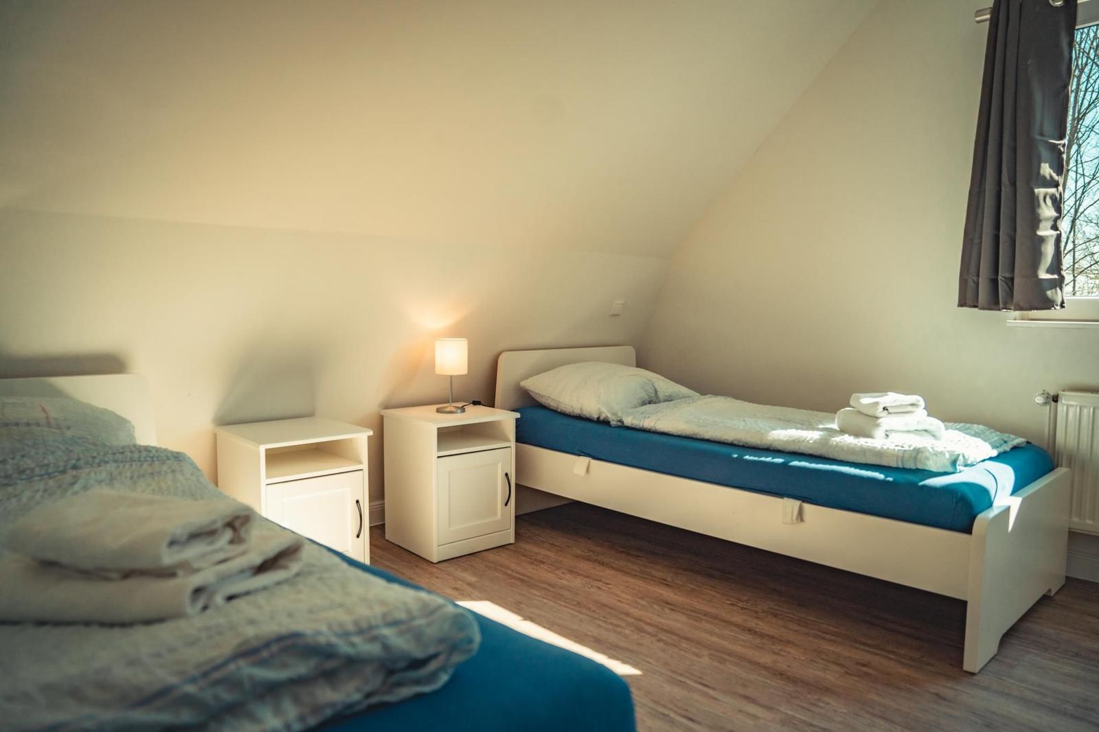 Zwei Betten mit blauen Matratzen und weißen Nachttischen. Fenster mit Vorhang.