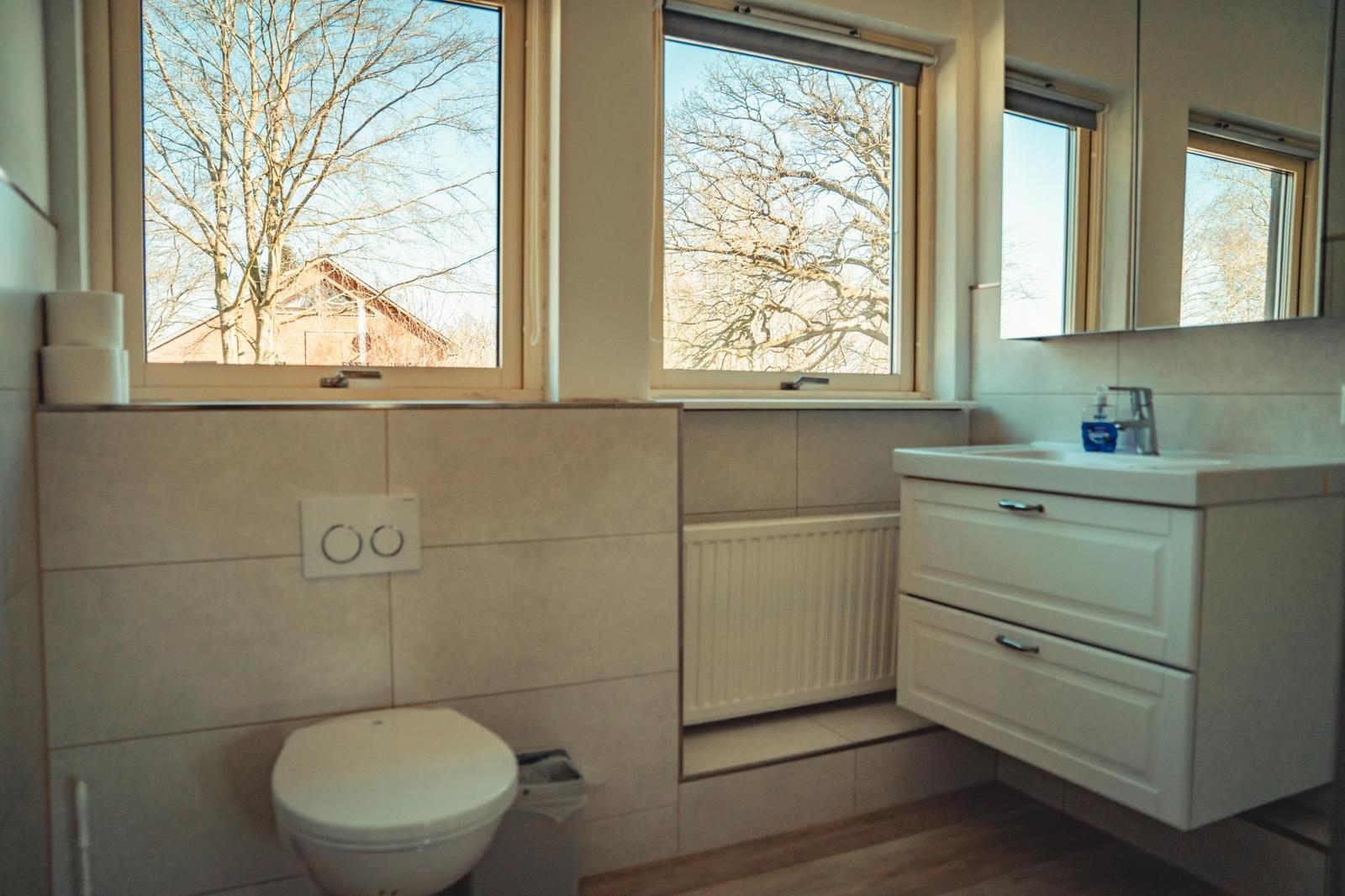 Badezimmer mit Toilette, Waschbecken und zwei Fenstern mit Blick auf Bäume.
