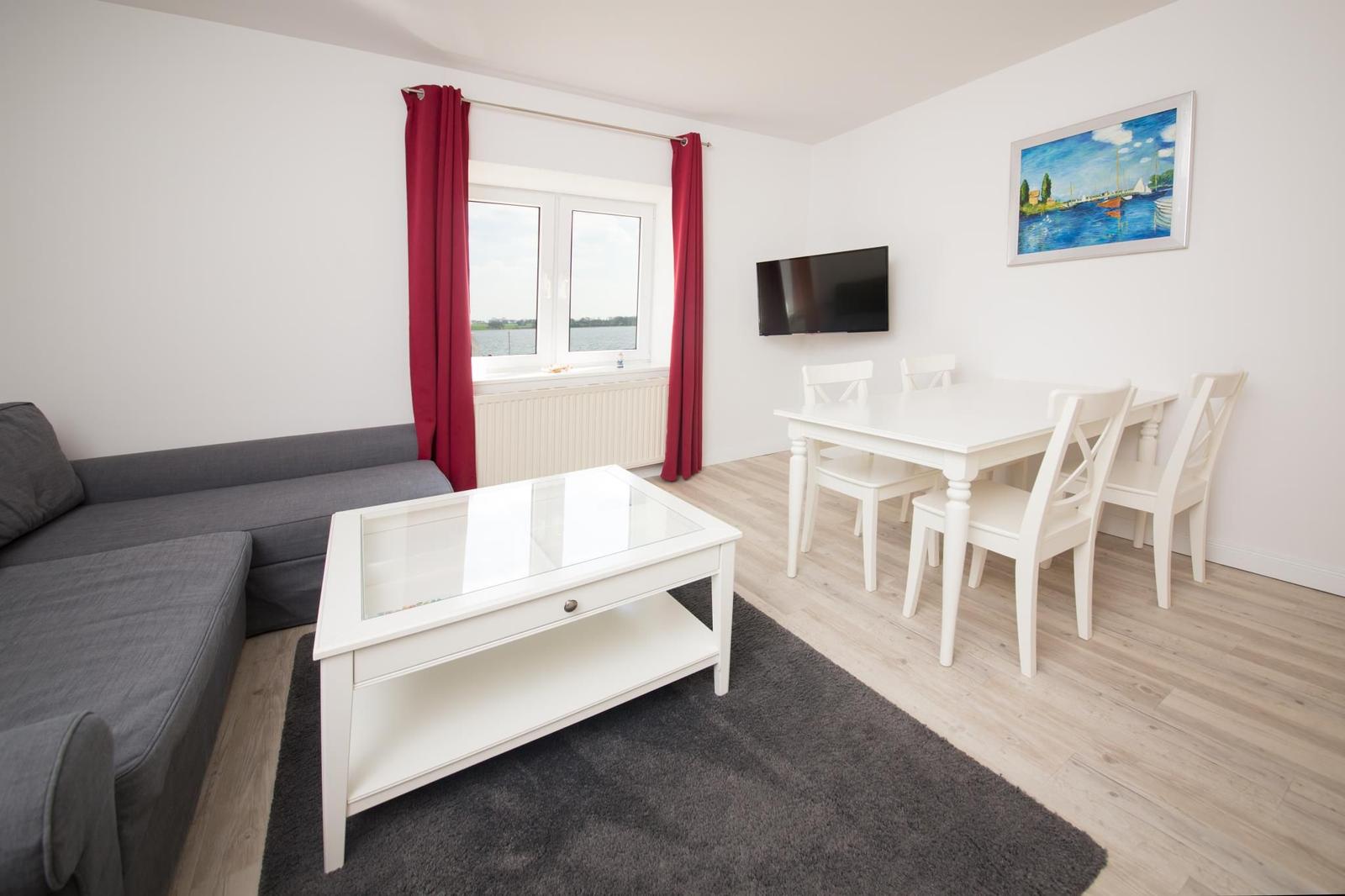 Wohn- und Essbereich mit Sofa, Tisch und Stühlen. Fenster mit roten Vorhängen. Wandbild und Fernseher.