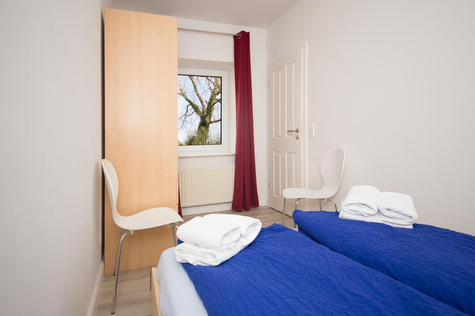 Zwei Betten mit blauen Bettdecken, weiße Stühle, Fenster mit rotem Vorhang.
