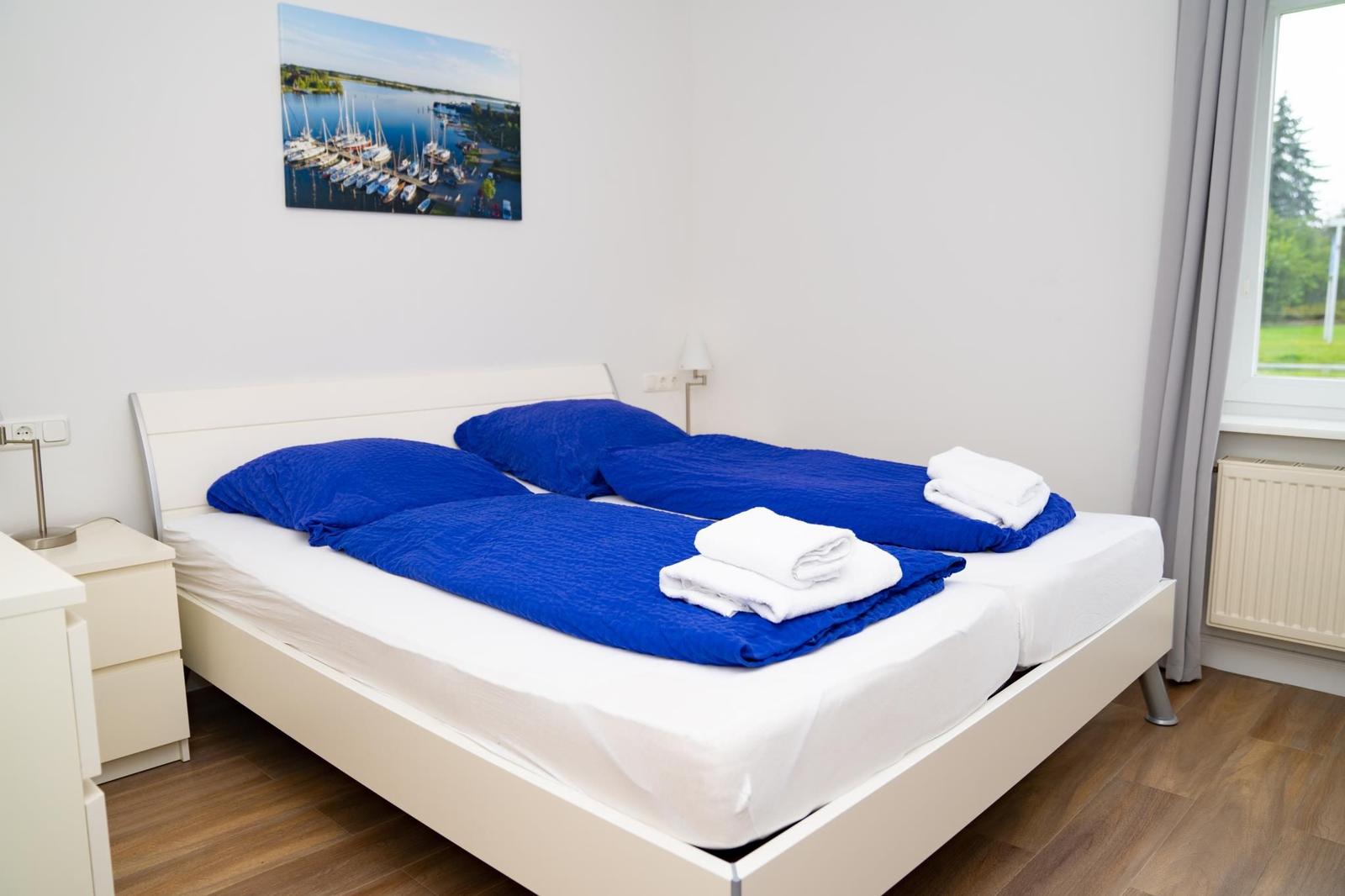 Schlafzimmer mit Doppelbett, blauen Bettwäsche, Fenster und Wandbild.