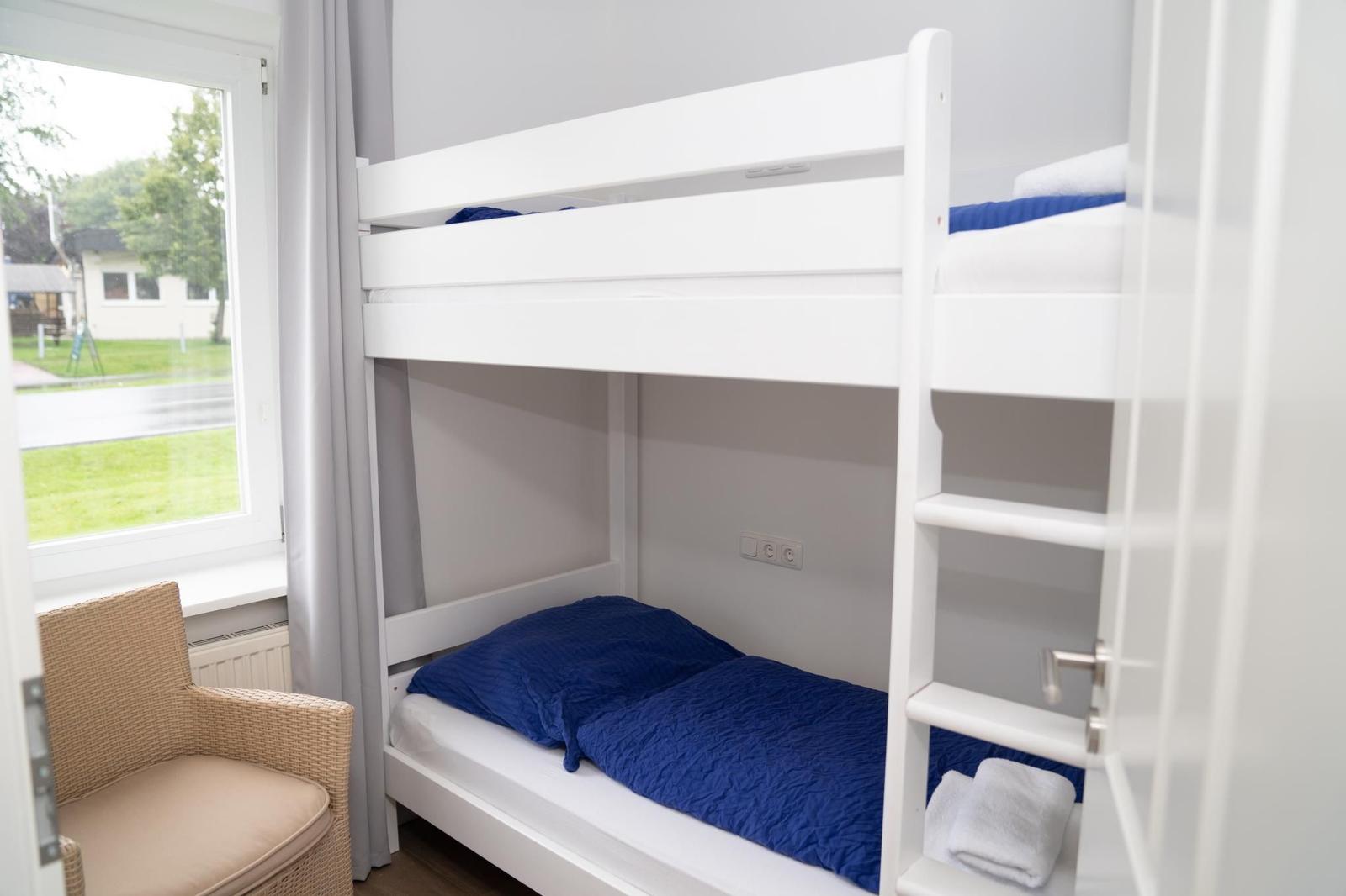 Zimmer mit weißen Doppelstockbetten, blauer Bettwäsche und Sitzgelegenheit.