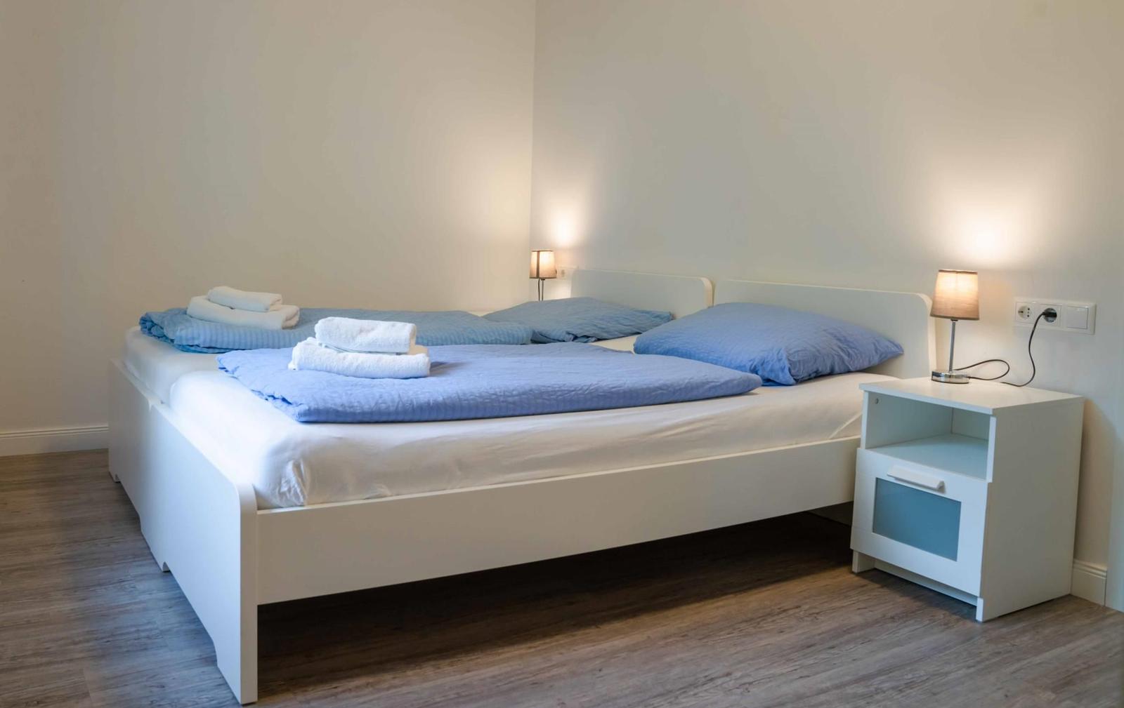 Schlafzimmer mit weißem Bett, blauen Kissen und Holzboden. Nachttisch mit Lampe rechts.