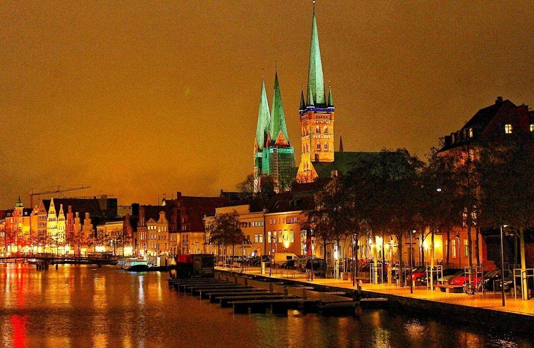 Night view of Lübeck harbor with prominent church towers and illuminated buildings along the water.