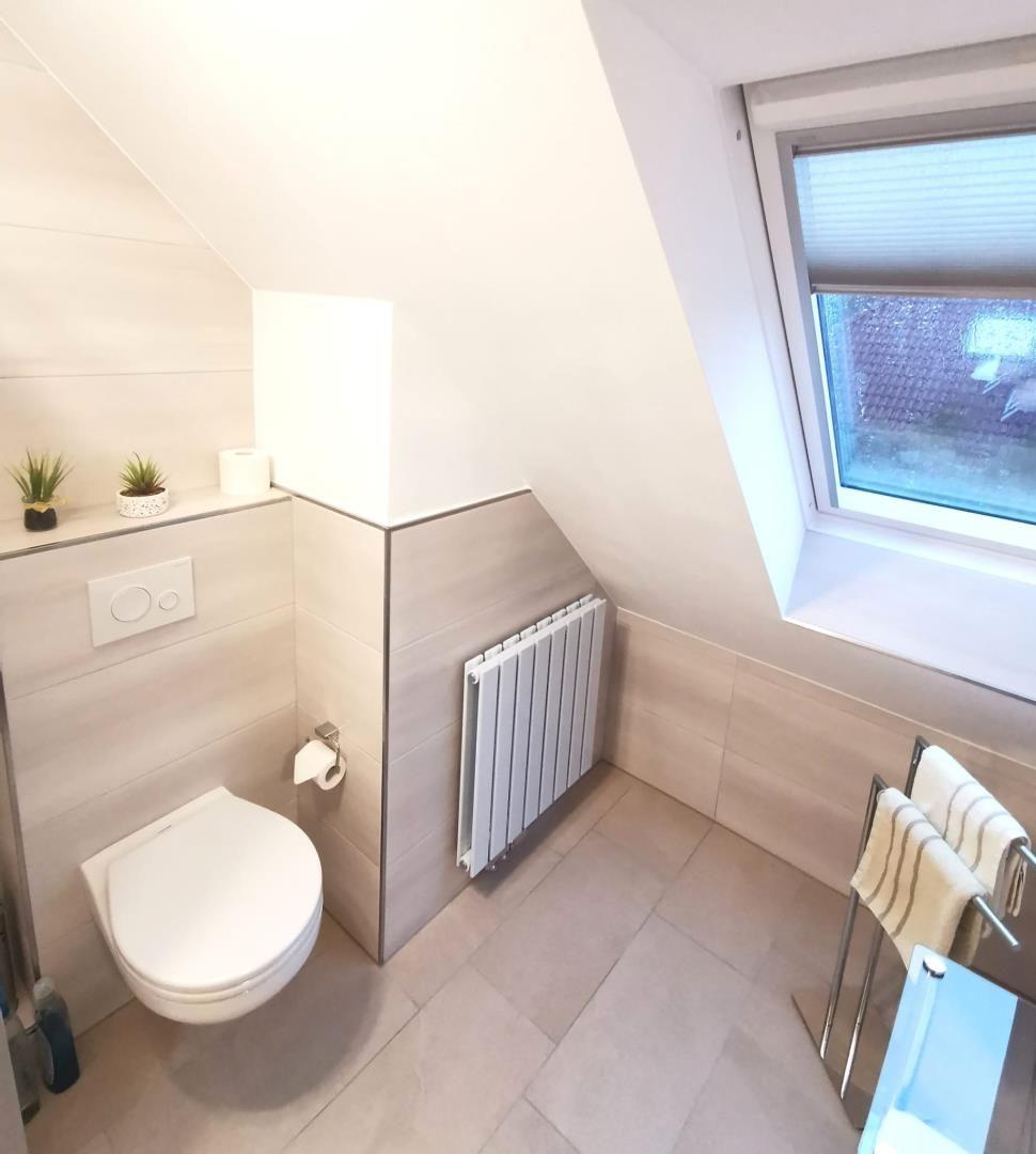 Badezimmer mit Toilette, Heizkörper und Fenster. Fliesen an Wänden und Boden.