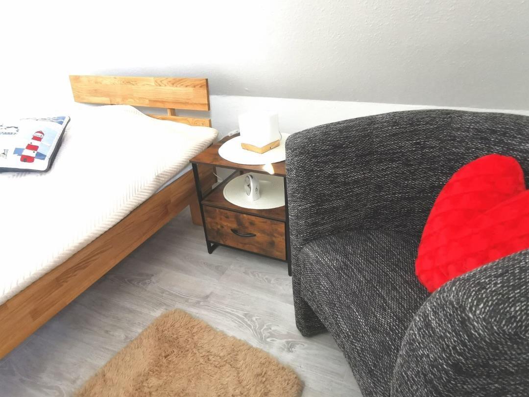Schlafzimmer mit Bett, Nachttisch und Sessel. Holzbett, grauer Sessel mit rotem Kissen.