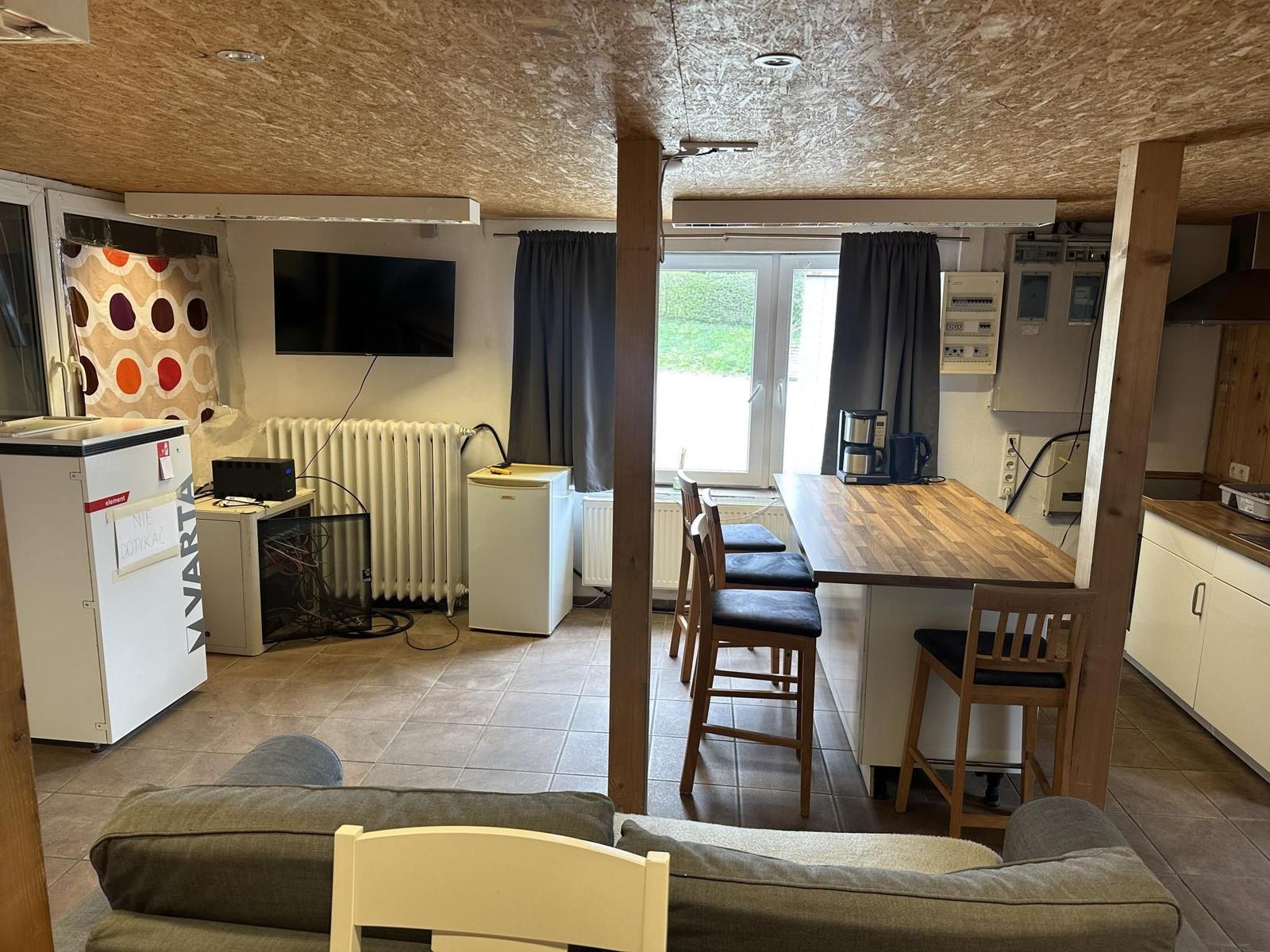 Wohn- und Küchenbereich mit Esstisch, Sofa und Fernseher. Holzdecke und Fenster mit Blick nach draußen.