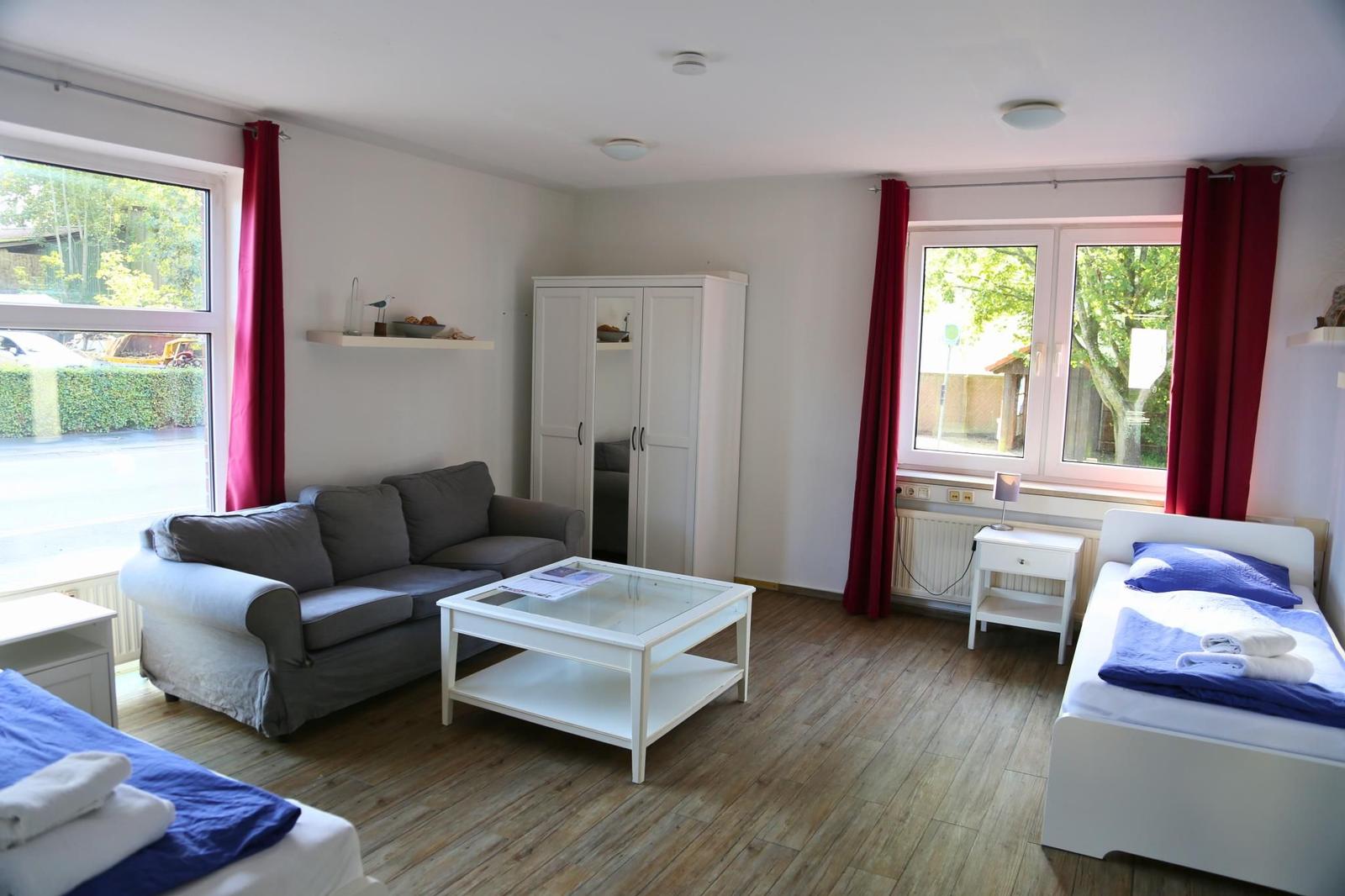 Wohnraum mit zwei Betten, Sofa, Schrank und zwei Fenstern mit roten Vorhängen.