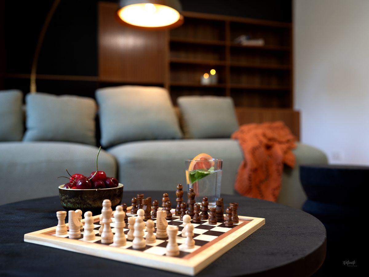 Schachbrett auf Tisch mit Kirschen und Wasser. Sofa mit Kissen und Holzregal im Hintergrund.