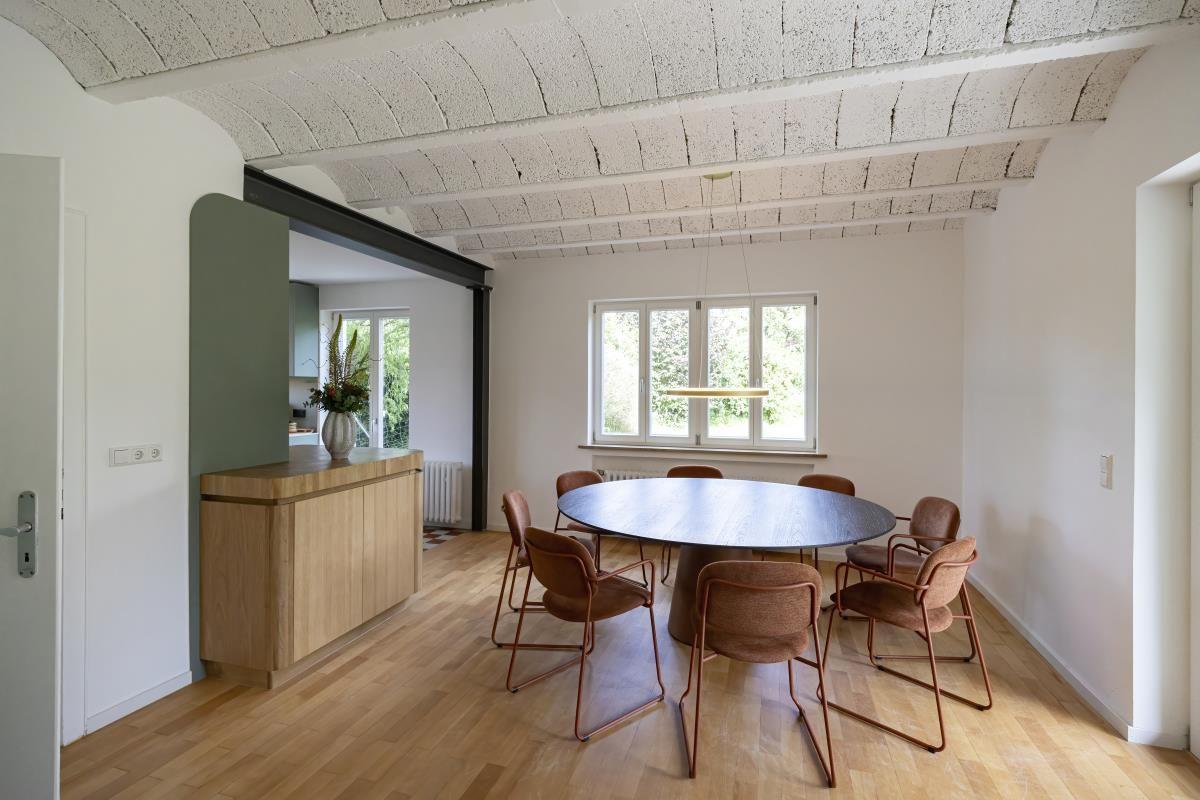 Esszimmer mit runden Tisch und Stühlen, Holzboden und Fenster. Küchenbereich im Hintergrund.