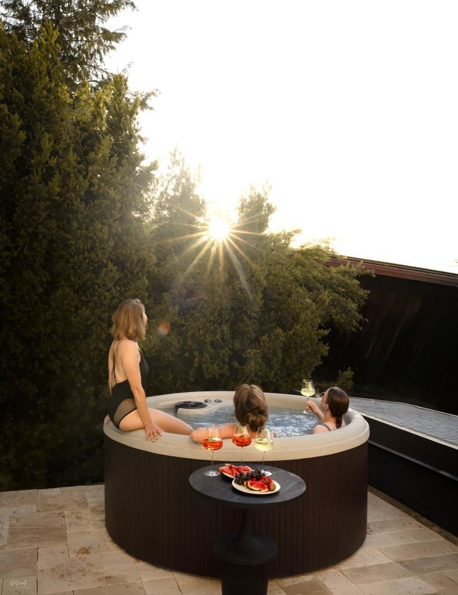 Drei Personen genießen einen Whirlpool auf einer Terrasse mit Blick auf Bäume und Sonnenuntergang.