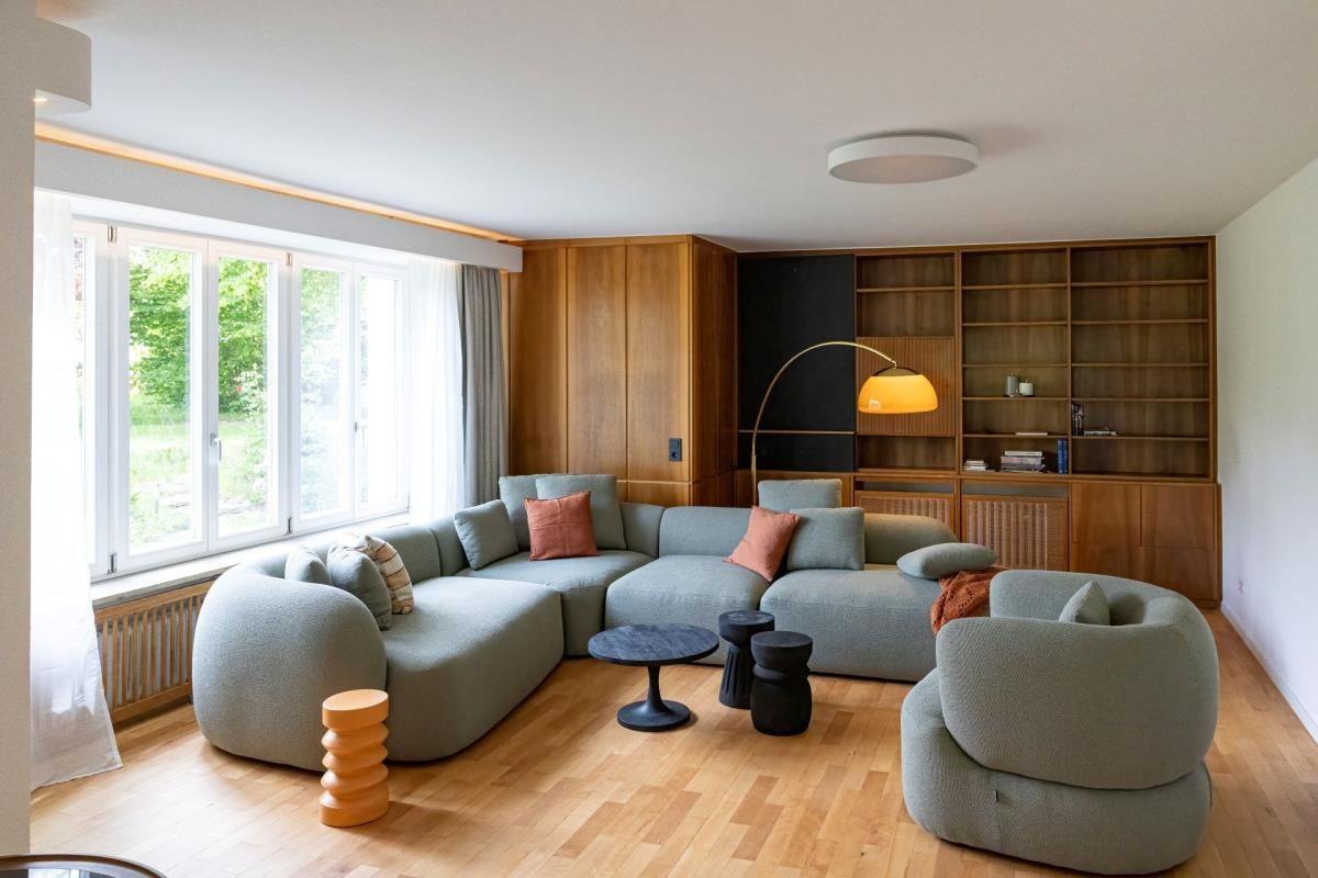 Wohnzimmer mit Sofa, Sessel und Holzmöbel. Große Fenster mit Blick auf Garten.
