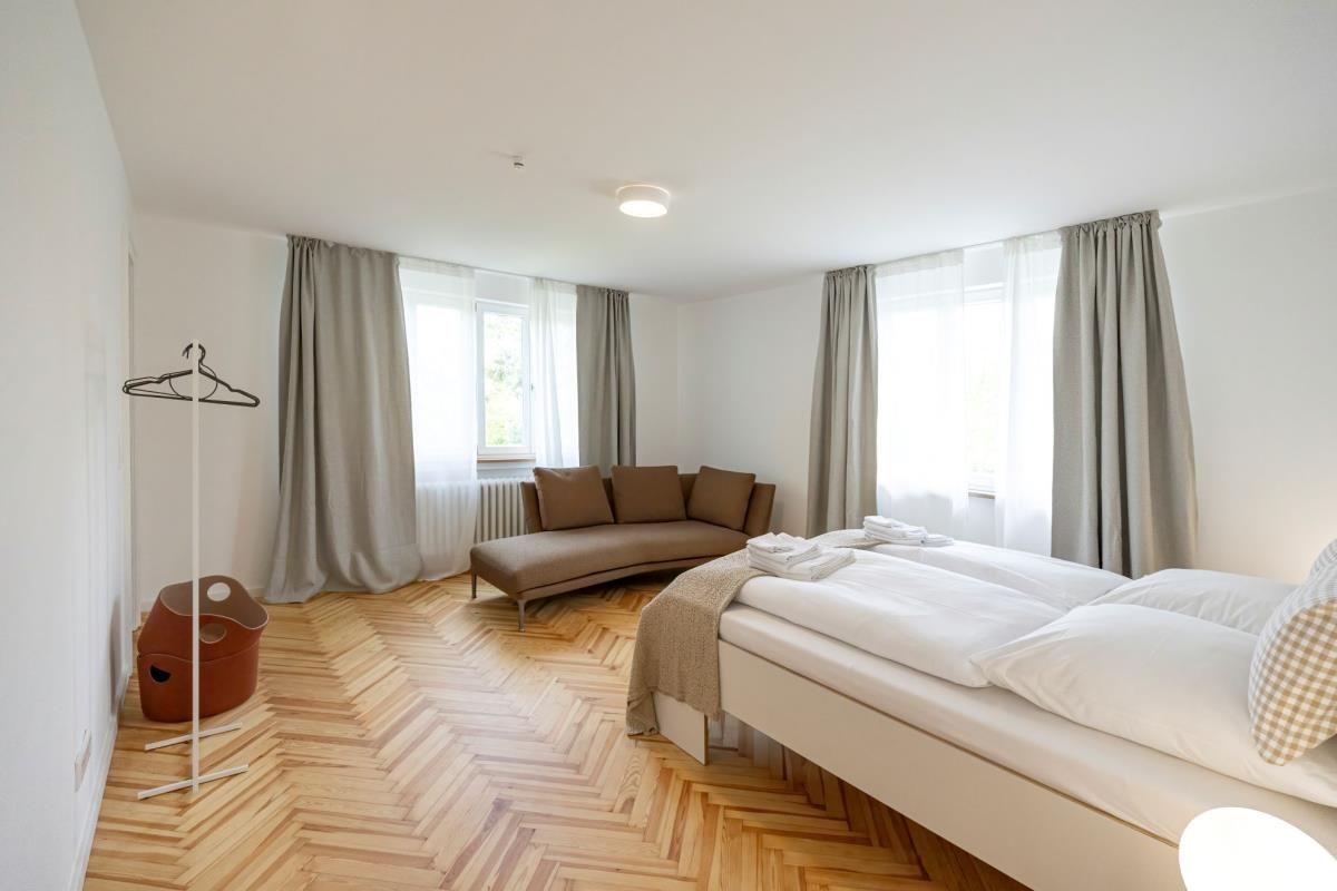 Schlafzimmer mit Bett, Sofa und Holzboden. Fenster mit Vorhängen.