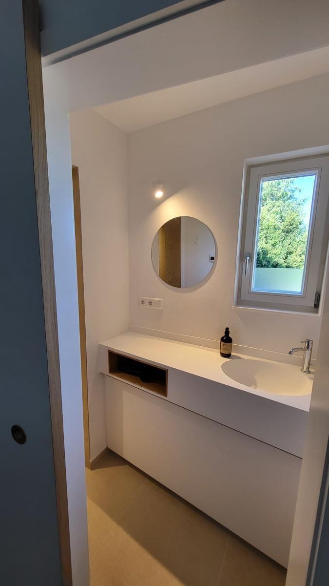 Badezimmer mit Waschbecken, runden Spiegel und Fenster mit Blick auf Bäume.