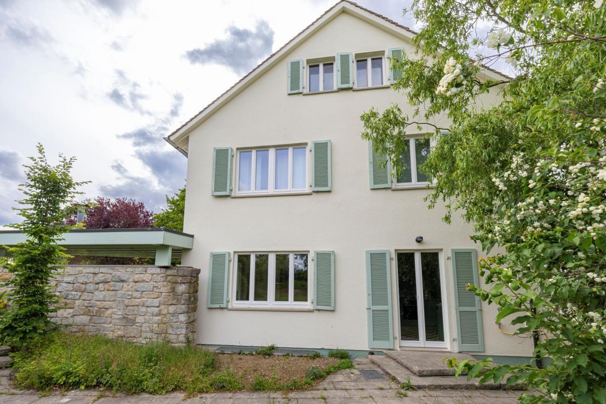 Ein zweistöckiges Haus mit hellen Wänden und grünen Fensterläden. Vor dem Haus wachsen Bäume und Sträucher.