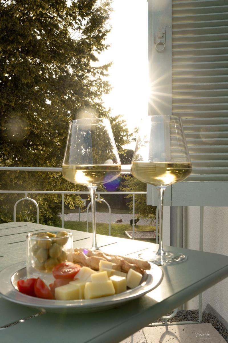 Zwei Weingläser und ein Teller mit Käse, Tomaten und Oliven auf einer Terrasse.