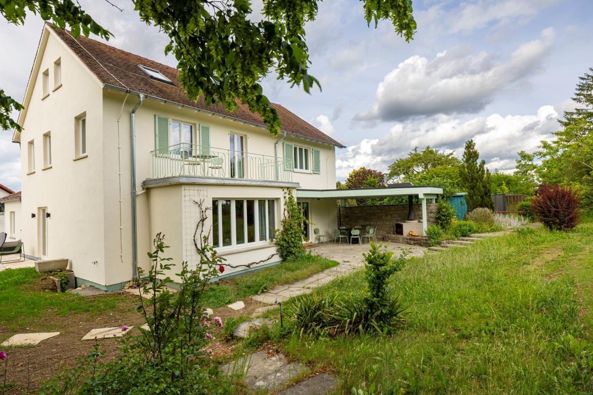 Ein zweistöckiges Haus mit Balkon, Garten und Terrasse unter einem bewölkten Himmel.