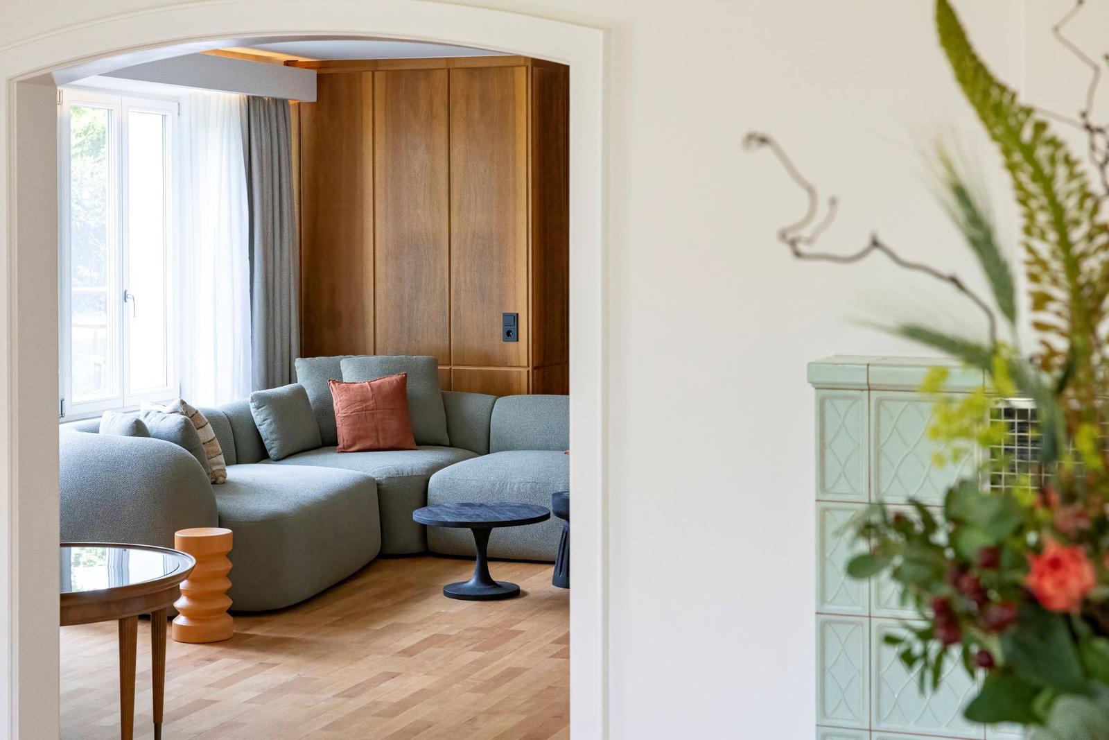Ein Wohnzimmer mit grauem Sofa, Holzboden und Holzwand. Fenster mit Vorhängen.