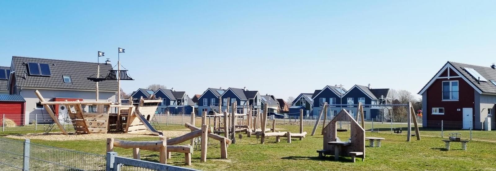 Großer Spielplatz mit Holzspielgeräten und Häusern im Hintergrund.