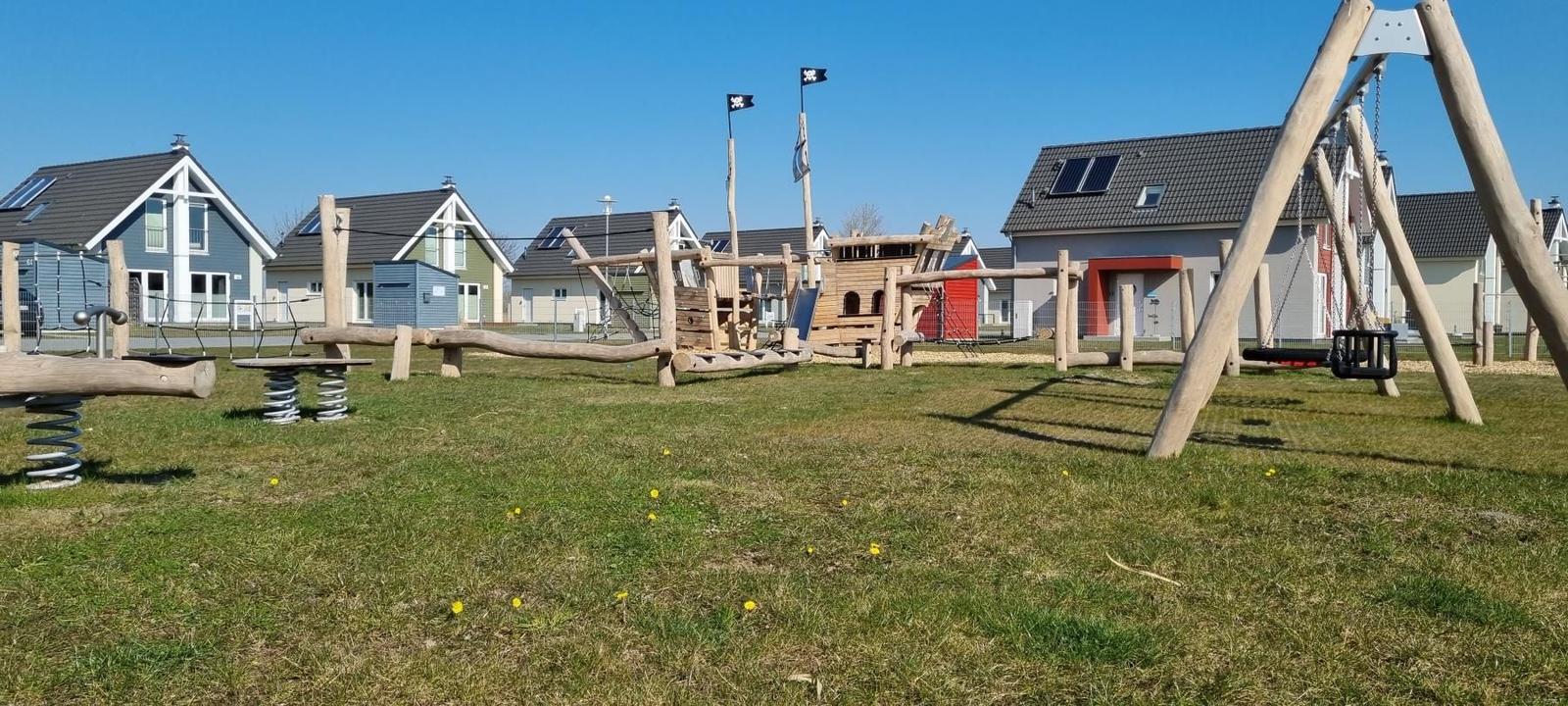 Großer Spielplatz mit Holzspielgeräten und Schaukeln vor modernen Häusern.